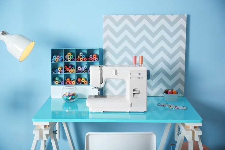 Sewing machine on a blue table with colorful threads, a chevron canvas, and a lamp in a well-lit room