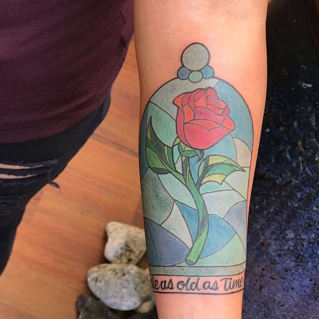 A Beauty and the Beast-inspired tattoo featuring a red rose inside a stained glass dome, with the quote "Tale as old as time" beneath it