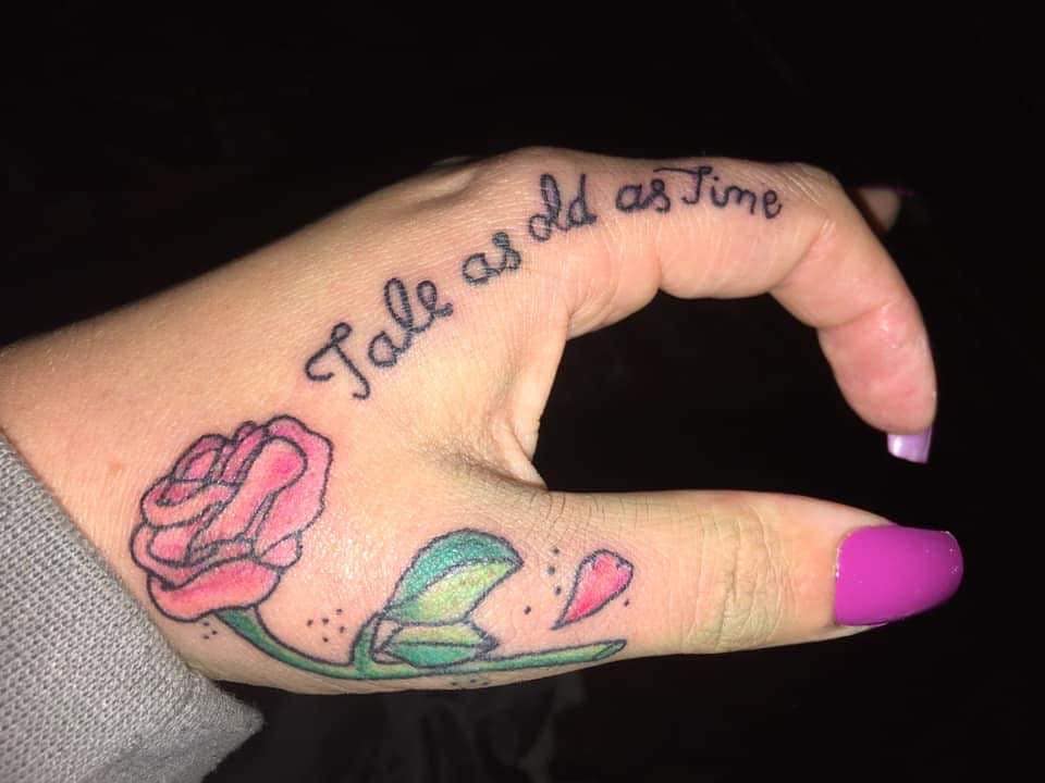 A hand tattoo featuring a pink rose with a green stem, accompanied by the quote "Tale as old as time" on the fingers