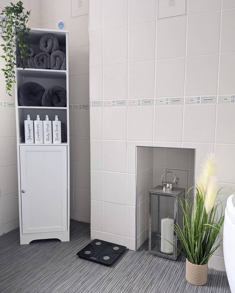 White bathroom cabinet with towels, labeled bottles, scale, and decorative plant.