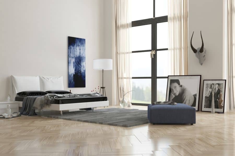 Luxury bedroom with tall windows, portrait art, blue ottoman, and neutral decor.