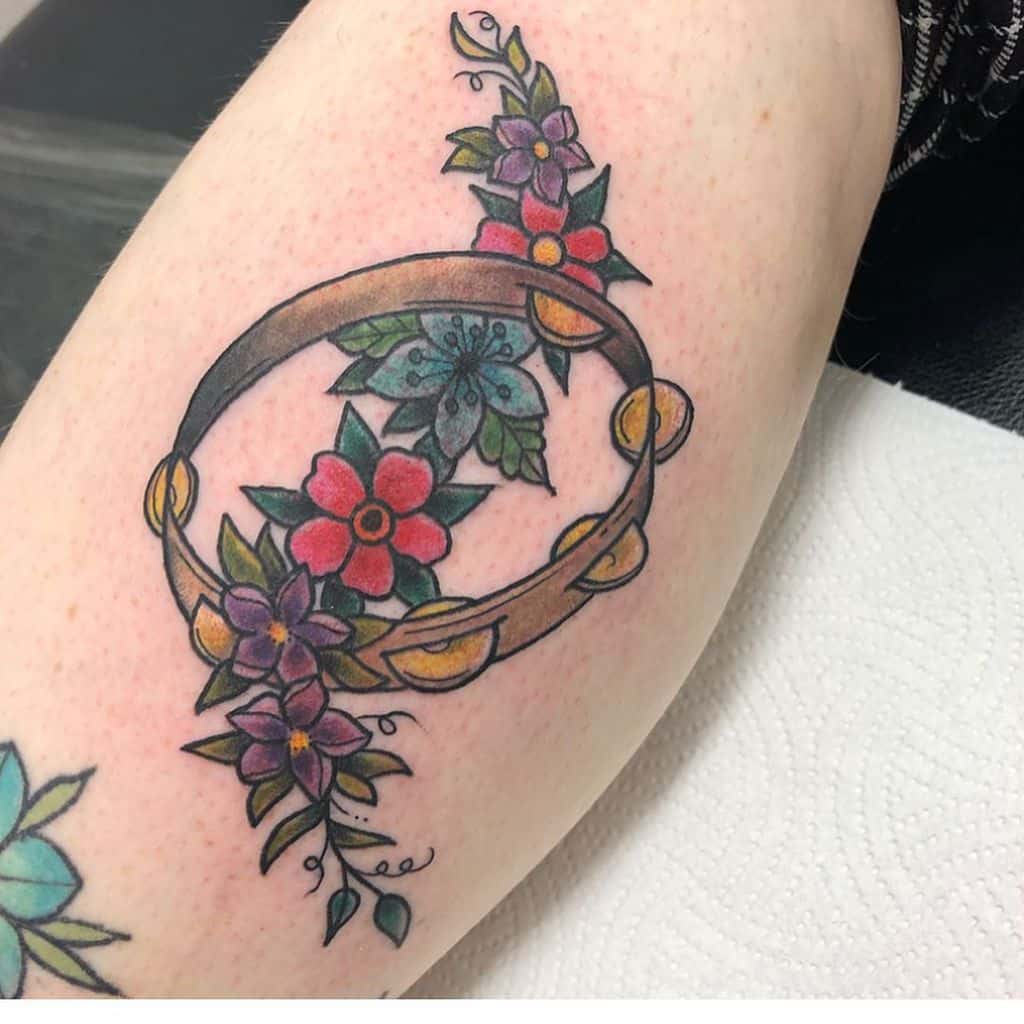Colorful tattoo of a tambourine wrapped in vibrant flowers and leaves, with gold jingles, on the upper arm.