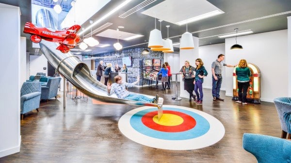 Office space with a slide, vibrant decor, socializing people, and features like a red plane sculpture and jukebox