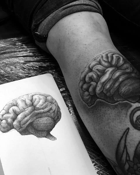 Black and grey tattoo of a human brain on the leg, closely matching a detailed dotwork-style sketch placed beside it.