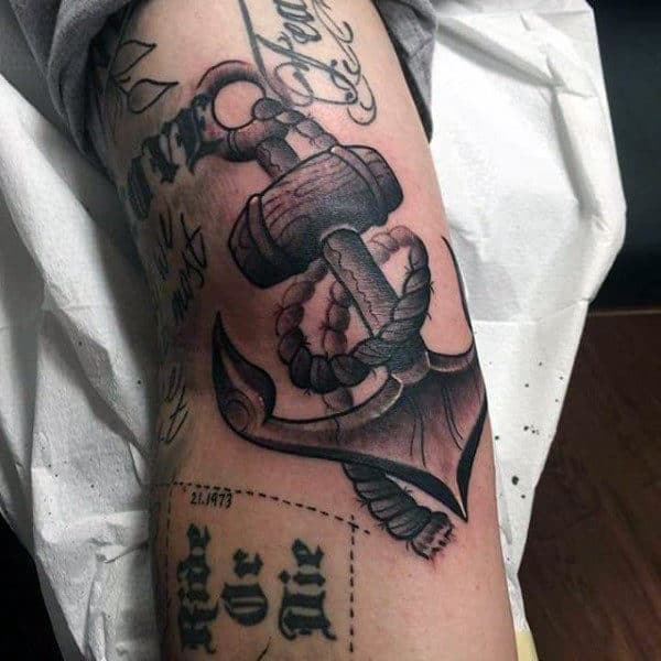 Tattoo of an anchor with rope on an arm, surrounded by writing and designs