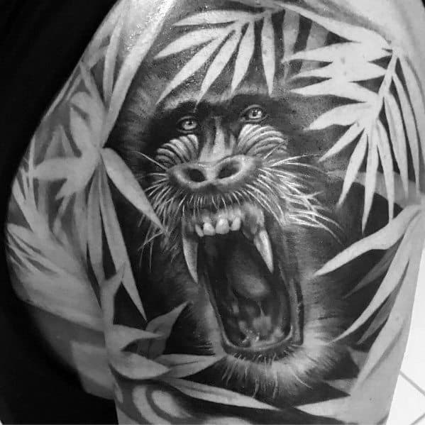 Black and grey realistic baboon tattoo with intense shading, fierce open-mouthed expression, and jungle foliage, creating a bold and dynamic shoulder design.