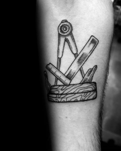 Black and gray tattoo of a compass, ruler, and chisel resting on a wood block, symbolizing precision, craftsmanship, and woodworking mastery.