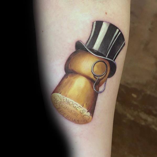 Tattoo of a champagne cork with a monocle and top hat, featuring "Venoge & Co, Champagne Epernay" on the cork, on a person's arm.
