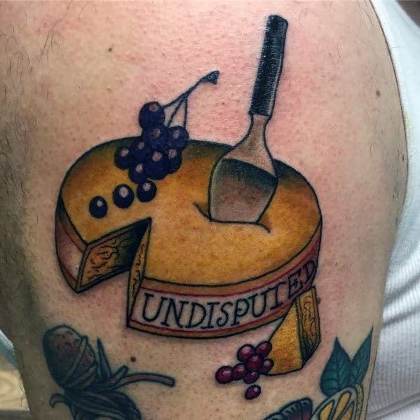 Colorful tattoo of a round cheese wheel with a slice cut out, topped with grapes and a cheese knife, featuring a ribbon that reads "UNDISPUTED" in bold lettering.