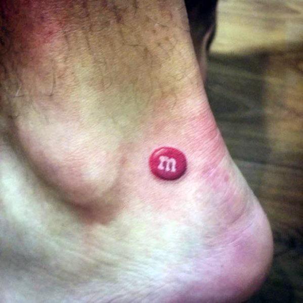 Realistic tattoo of a small red M&M candy on the ankle, featuring detailed shading and highlights for a 3D effect.