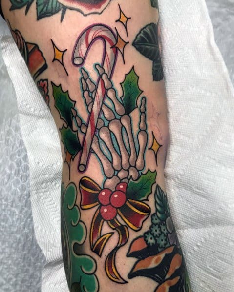 Christmas tattoo featuring a skeletal hand holding a candy cane, surrounded by holly leaves, berries, and a festive ribbon for a spooky holiday twist.