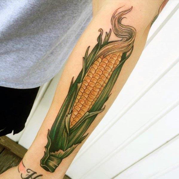 Bold tattoo of a detailed corn cob with green husks and golden kernels, stretching along the forearm with vibrant shading.
