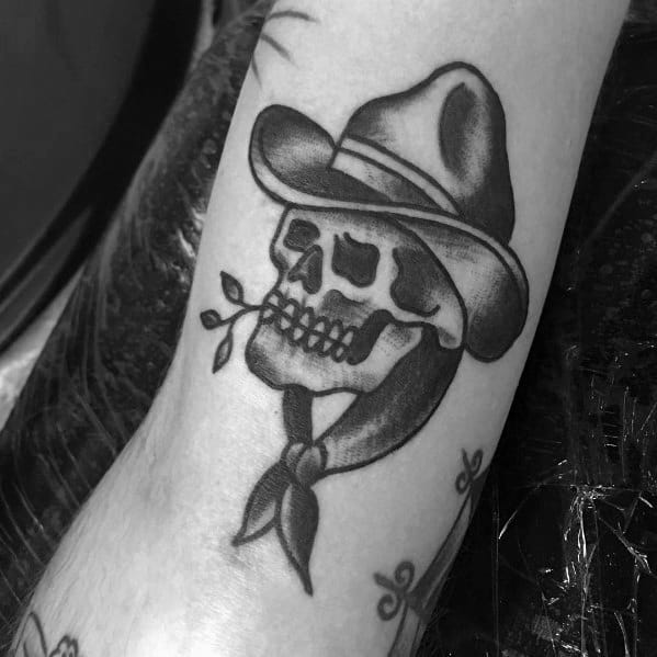 Black and gray tattoo of a cowboy skull wearing a hat and bandana, holding a twig in its teeth, done in a bold traditional Western style.