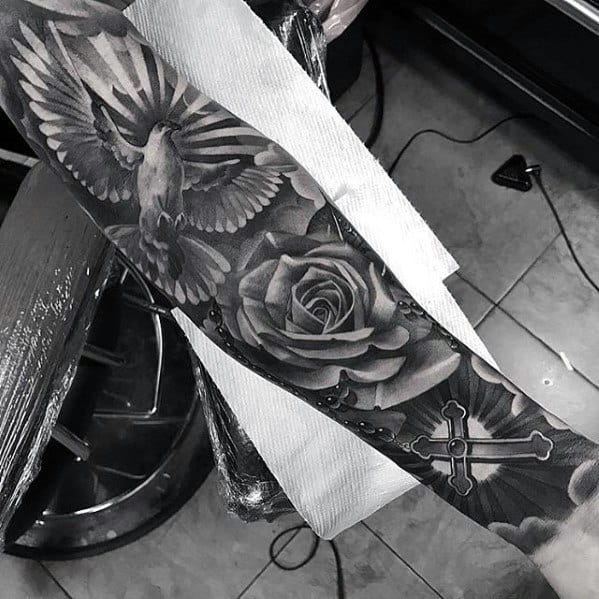 A stunning black and gray sleeve tattoo featuring a dove, rose, and cross adorned with beads elegantly wraps around the arm