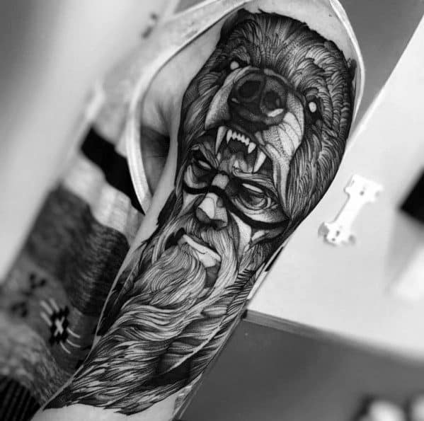 Black and grey upper arm tattoo featuring a warrior with a fierce expression, wearing a roaring bear headdress for a bold and powerful look.
