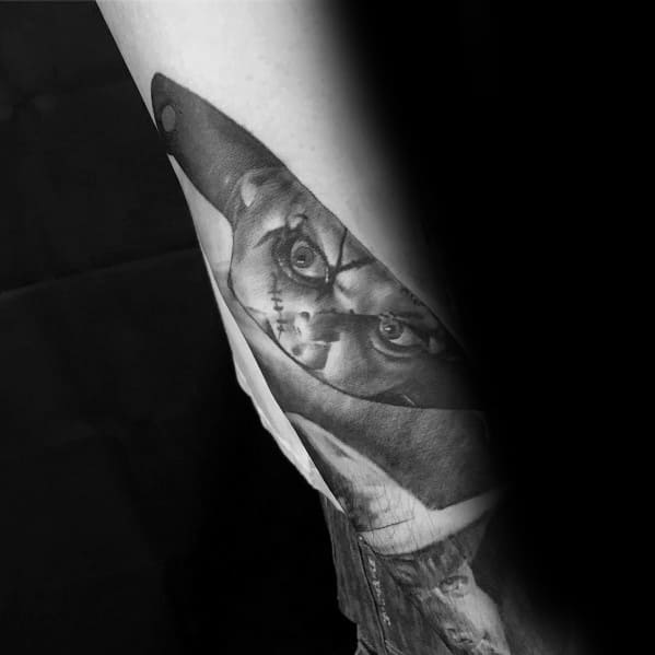 Black and grey Chucky tattoo with his menacing face reflected in a realistic knife blade, creating a chilling and creative horror-inspired design.