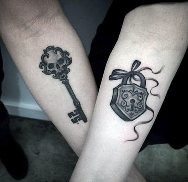 Matching forearm tattoos of an ornate key with a skull design and a heart-shaped lock with a ribbon, symbolizing a dark yet deep bond and being each other’s perfect match.