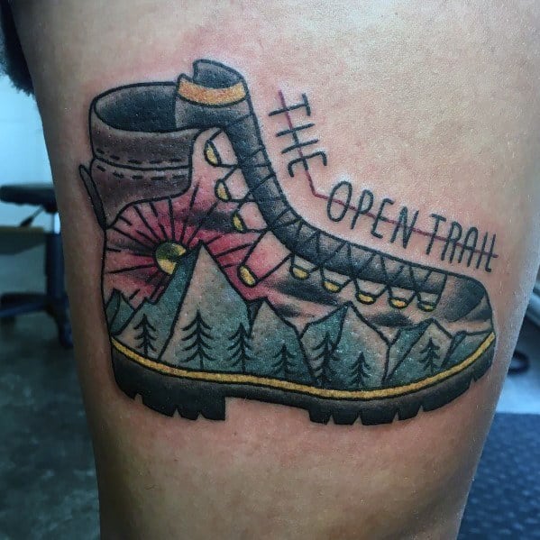 A tattoo of a hiking boot with a mountain landscape inside, featuring pine trees, a sunset, and "The Open Trail" written along the boot.