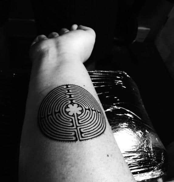 Circular labyrinth tattoo on a man's forearm, featuring a bold maze design with a four-leaf clover at the center and a gear-like outer edge.