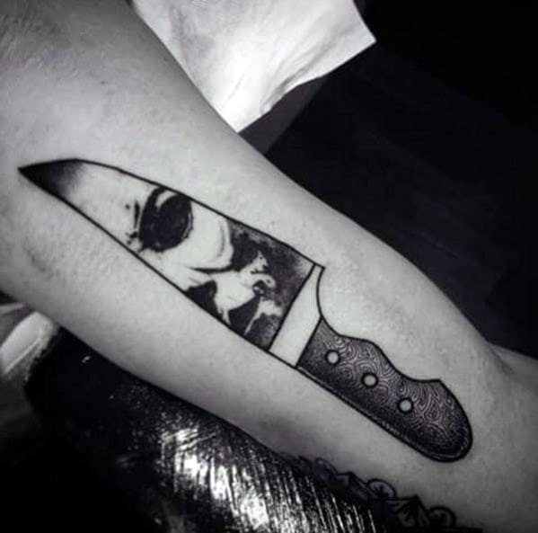 Black and grey tattoo of a knife with Michael Myers' eerie reflection in the blade, creating a chilling horror-inspired design.