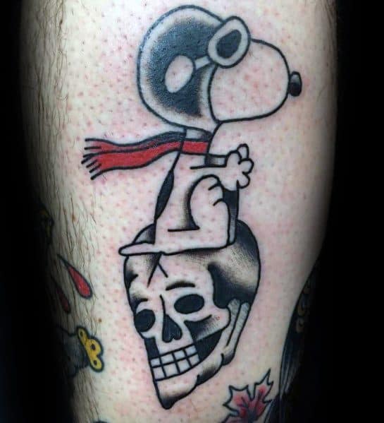 Colorful tattoo of Snoopy as a flying ace, wearing goggles and a red scarf, sitting on a detailed skull, placed on a person's leg.