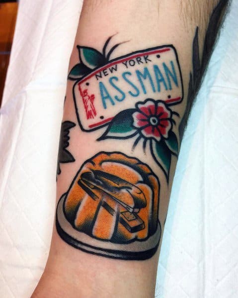 Color tattoo featuring a stapler encased in Jell-O, referencing Jim’s prank on Dwight from *The Office*, alongside a New York "ASSMAN" license plate from *Seinfeld*.