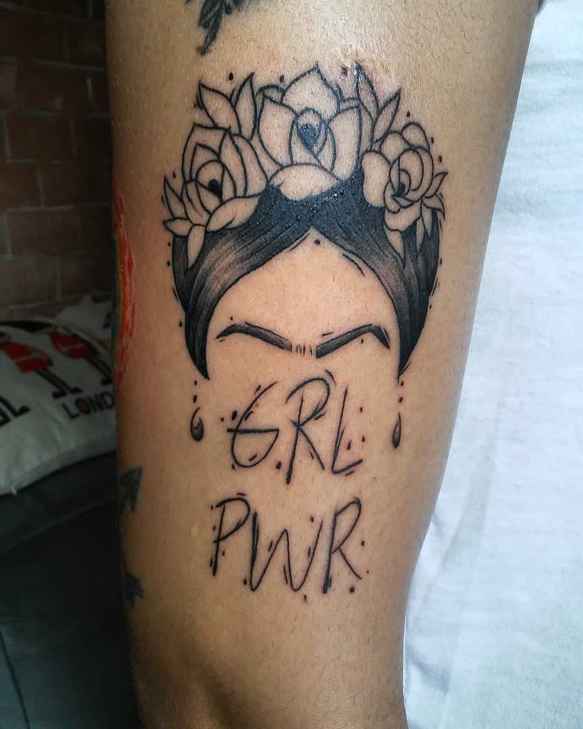 Black ink tattoo of a minimalist Frida Kahlo-inspired face with a floral crown, accompanied by the words 'GRL PWR' in bold lettering
