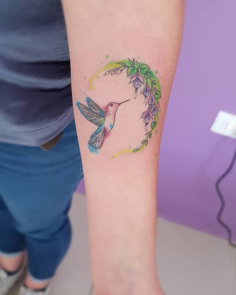 Vibrant watercolor-style tattoo of a hummingbird hovering near a crescent-shaped floral vine with delicate purple flowers and green leaves