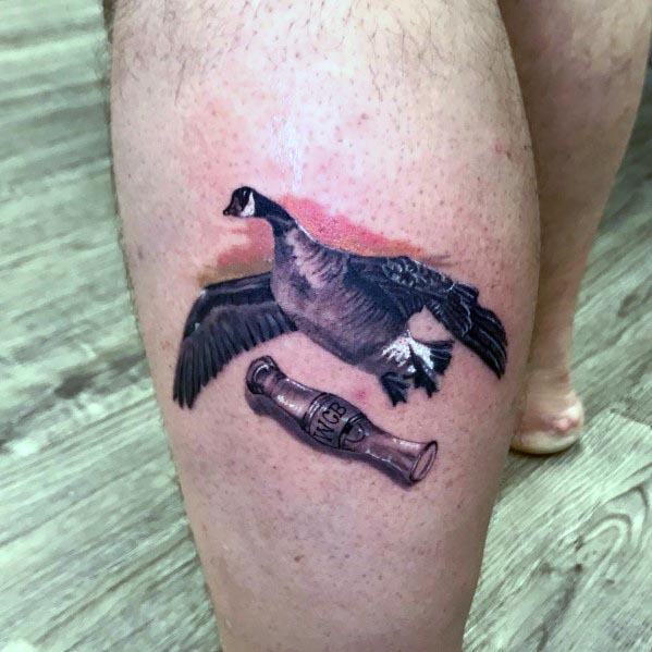 Realistic tattoo of a flying goose with a detailed beer bottle labeled "BAM" beneath it, inked on the calf with fine shading and color accents.