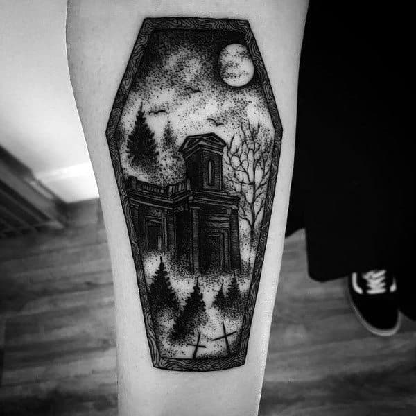 Black and grey tattoo featuring an eerie abandoned house in a coffin-shaped frame, surrounded by trees, a graveyard, and a full moon for a gothic look.
