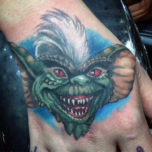 Gremlin hand tattoo with fierce red eyes, sharp teeth, and white hair, featuring bold colors and a vibrant blue background.