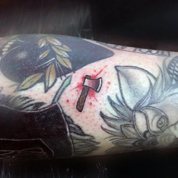 Tiny hatchet tattoo with a red-tipped blade on a heavily tattooed arm, featuring bold shading and a subtle red splatter effect for added impact.