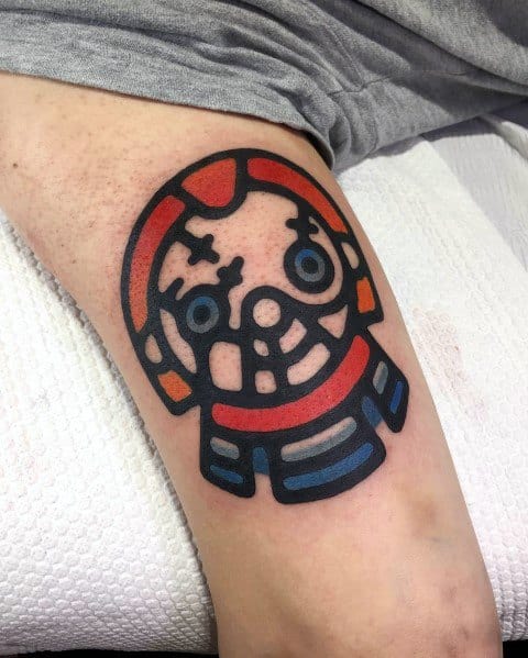 Minimalist Chucky tattoo with a bold, cartoon-style design, featuring his stitched face, bright blue eyes, and signature overalls for a unique look.
