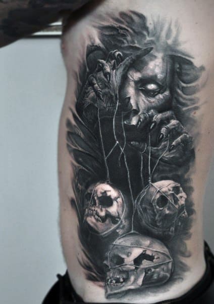 Black and grey rib tattoo featuring a sinister puppet master controlling skulls with strings, creating a dark and eerie gothic aesthetic.