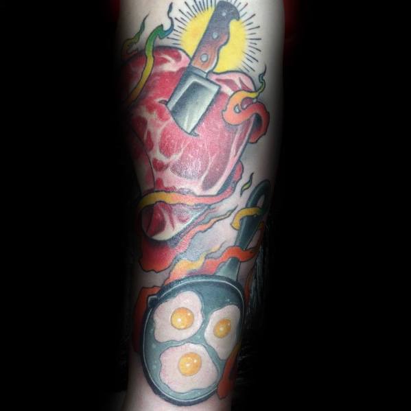 Tattoo of a raw steak pierced by a cleaver, surrounded by flames, with a frying pan of eggs below, creating a bold and dynamic food-inspired design.