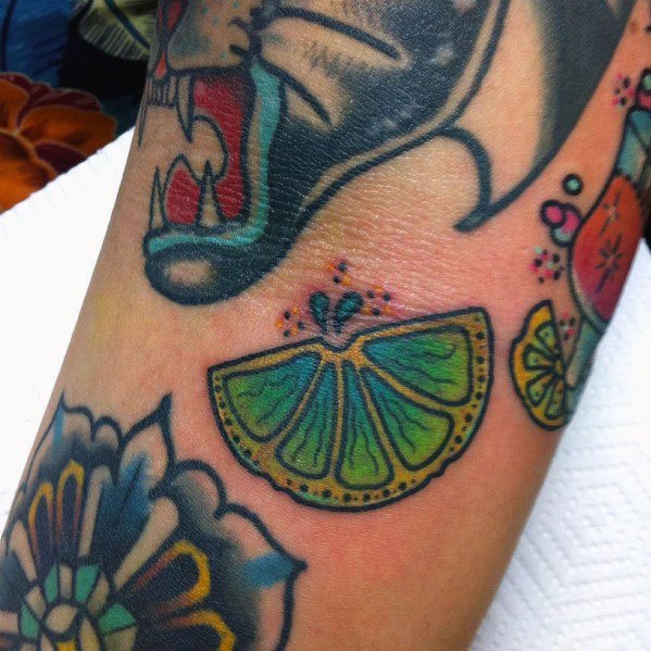 Traditional-style lime wedge tattoo with bold outlines, vibrant green shading, and decorative details, complementing an eclectic tattoo collection.