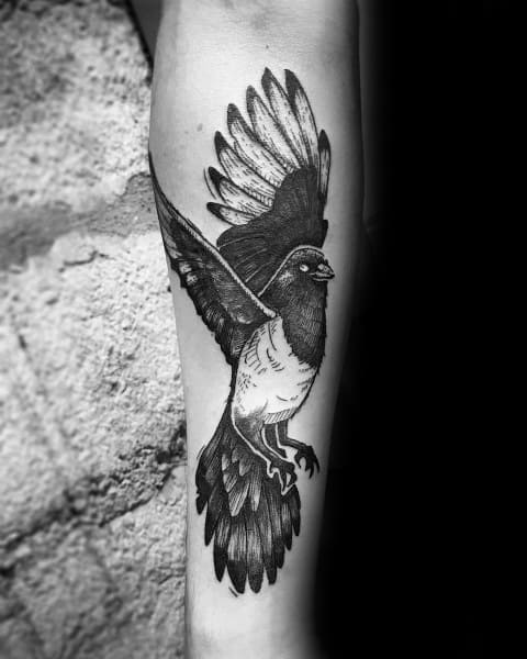 Black and gray magpie tattoo on forearm, featuring the bird in mid-flight with detailed feathers and bold shading for a striking look.
