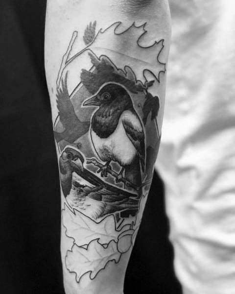Black and white tattoo of two magpies perched on a branch, surrounded by oak leaves, on a person's arm.