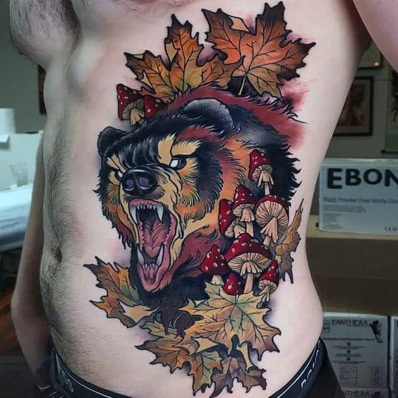 Neo-traditional bear tattoo on the side torso featuring a roaring bear surrounded by autumn leaves, red mushrooms, and bold shading for depth.