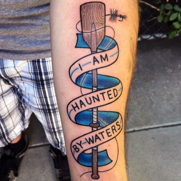 Colorful tattoo of a wooden paddle wrapped in a blue ribbon with the phrase "I AM HAUNTED BY WATERS," symbolizing a deep connection to water and adventure.