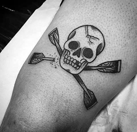 Black and grey tattoo of a skull with crossed paddles, resembling a pirate emblem, symbolizing adventure, resilience, and a fearless spirit on the water.