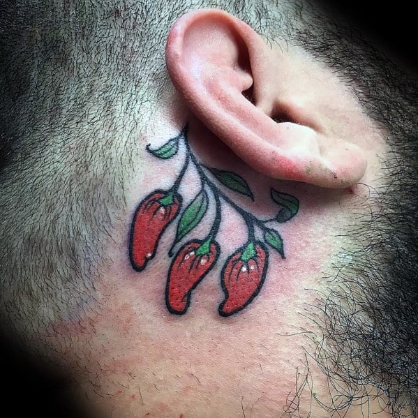 Small chili pepper tattoo behind the ear, featuring three red peppers with green leaves and fine black outlines for a delicate yet bold design.