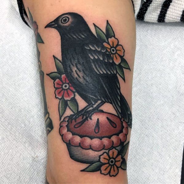 Traditional-style tattoo featuring a blackbird perched on a pie, surrounded by flowers, blending classic Americana with a whimsical folk tale vibe
