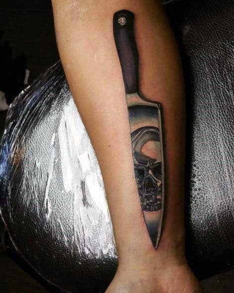 Tattoo of a knife with a skull reflection in the blade, creating a bold and eerie design with detailed shading and realism.
