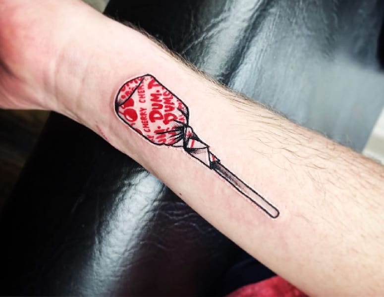 Minimalist Dum Dums lollipop tattoo with fine line details and subtle shading, creating a simple yet realistic design on the forearm.