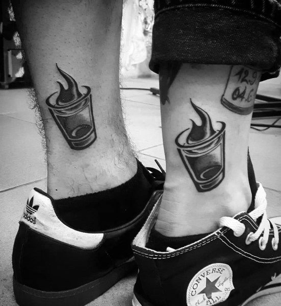 Matching black and grey flaming shot glass tattoos on the ankles, symbolizing boldness and celebration with a sleek and dynamic design.