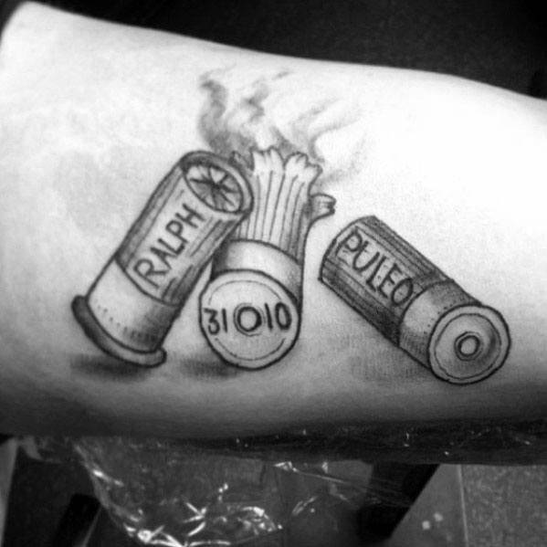 Black and grey tattoo of smoking shotgun shells with personalized names and dates, featuring fine shading and intricate detailing for a bold tribute design.