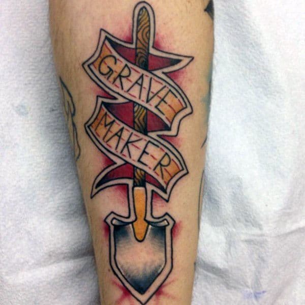 Shovel tattoo with a ribbon wrapped around it reading "Grave Maker," featuring bold colors and shading, symbolizing mortality and hard work.