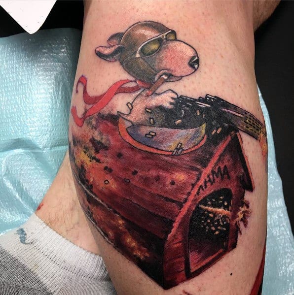 Colorful tattoo of Snoopy as a flying ace, wearing goggles and a red scarf, firing a machine gun while riding his battle-damaged doghouse.