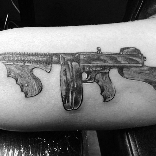 Realistic black and grey Tommy gun tattoo on the forearm, featuring intricate shading and wood grain details for a bold and detailed firearm design.